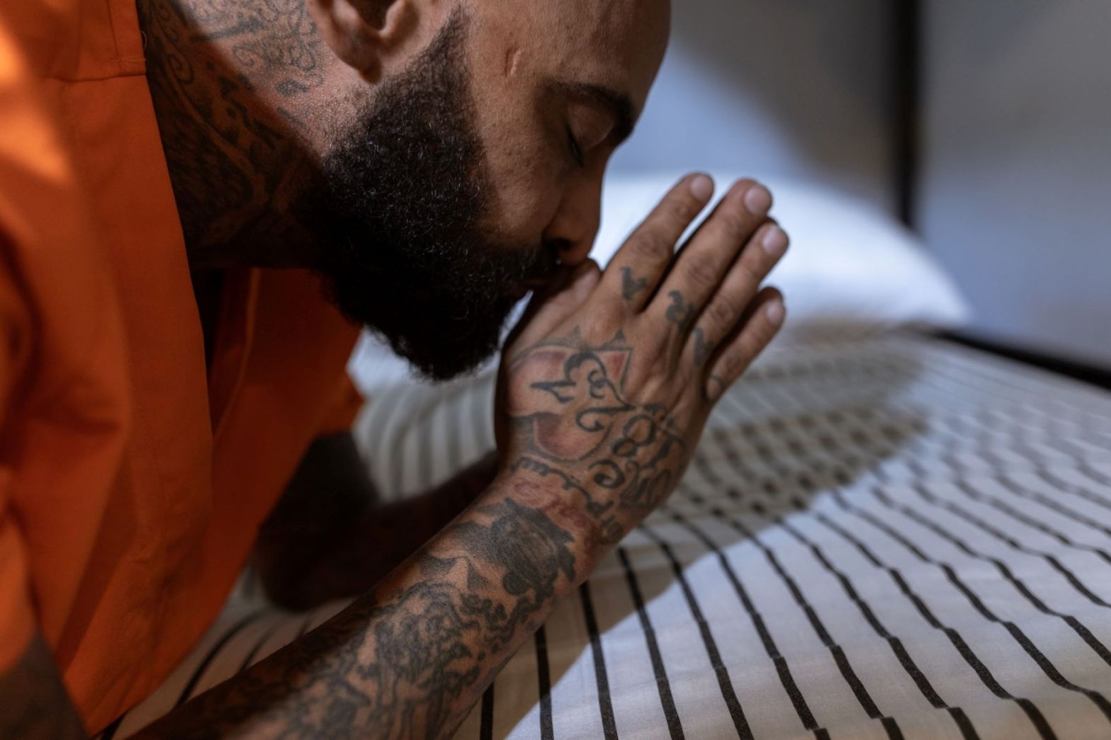 A tattooed convict clasps his hand in prayer beside his cell bed.